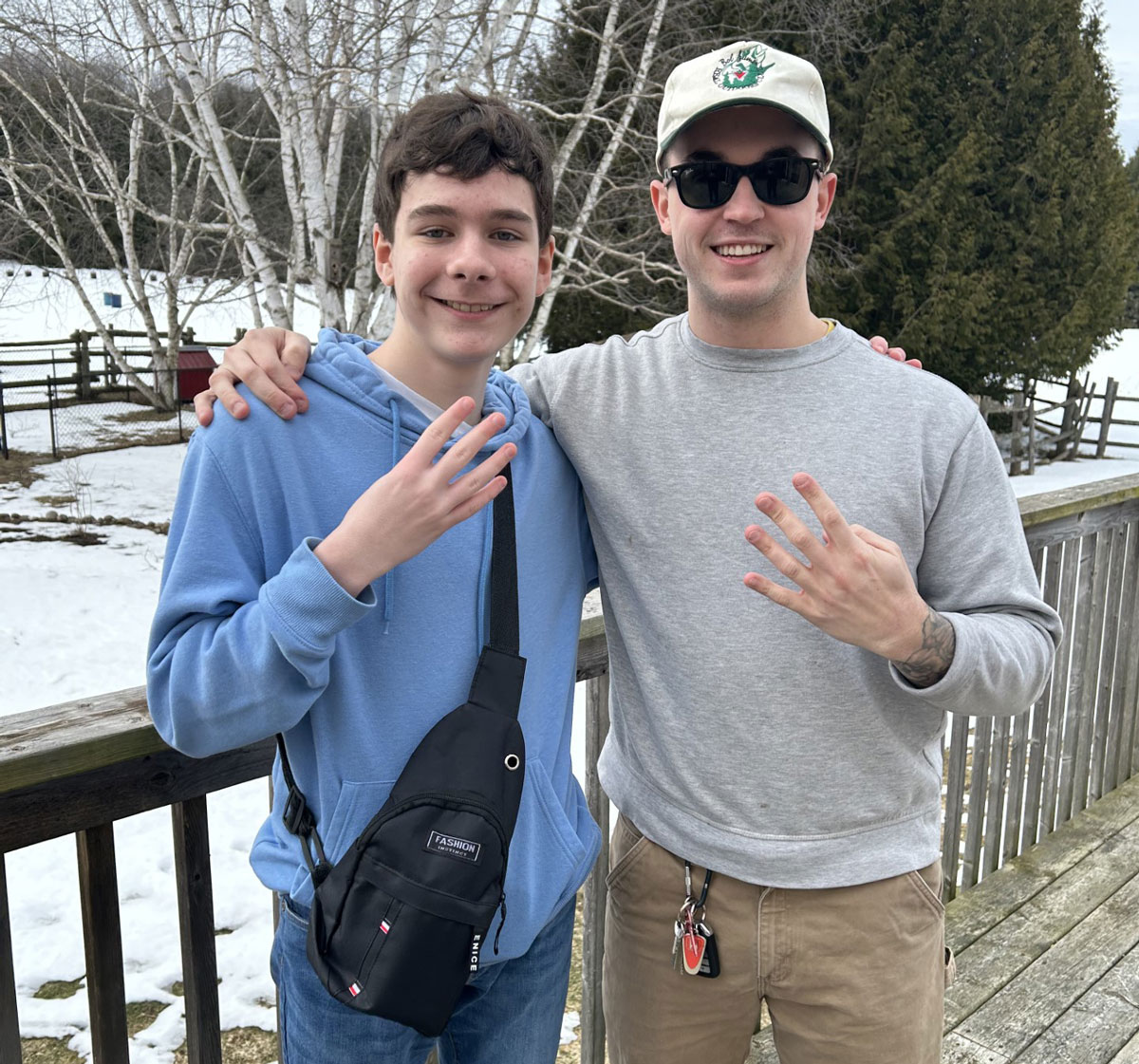 An older teen in a blue sweatshirt and a man in a grey sweatshirt and white ball cap have their arms around each other and are holding up 4 fingers.