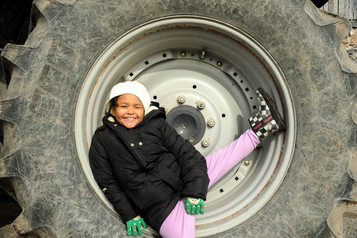 A young girl in a black jacket and pink pants smiles as she sits inside the rim of a huge tractor tire.