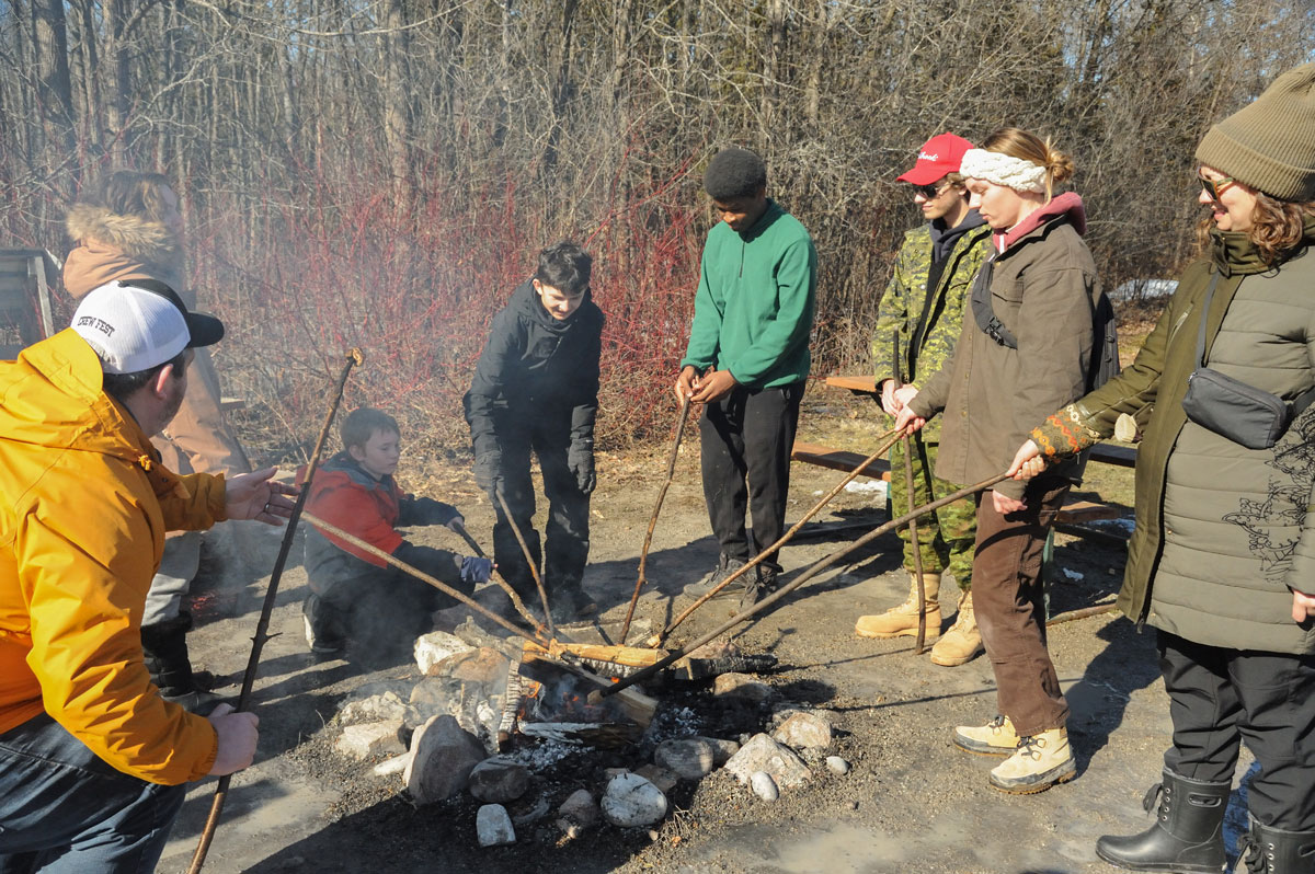 Adults, teens and a boy are standing around an open fire roasting marshmallows on long wooden sticks.