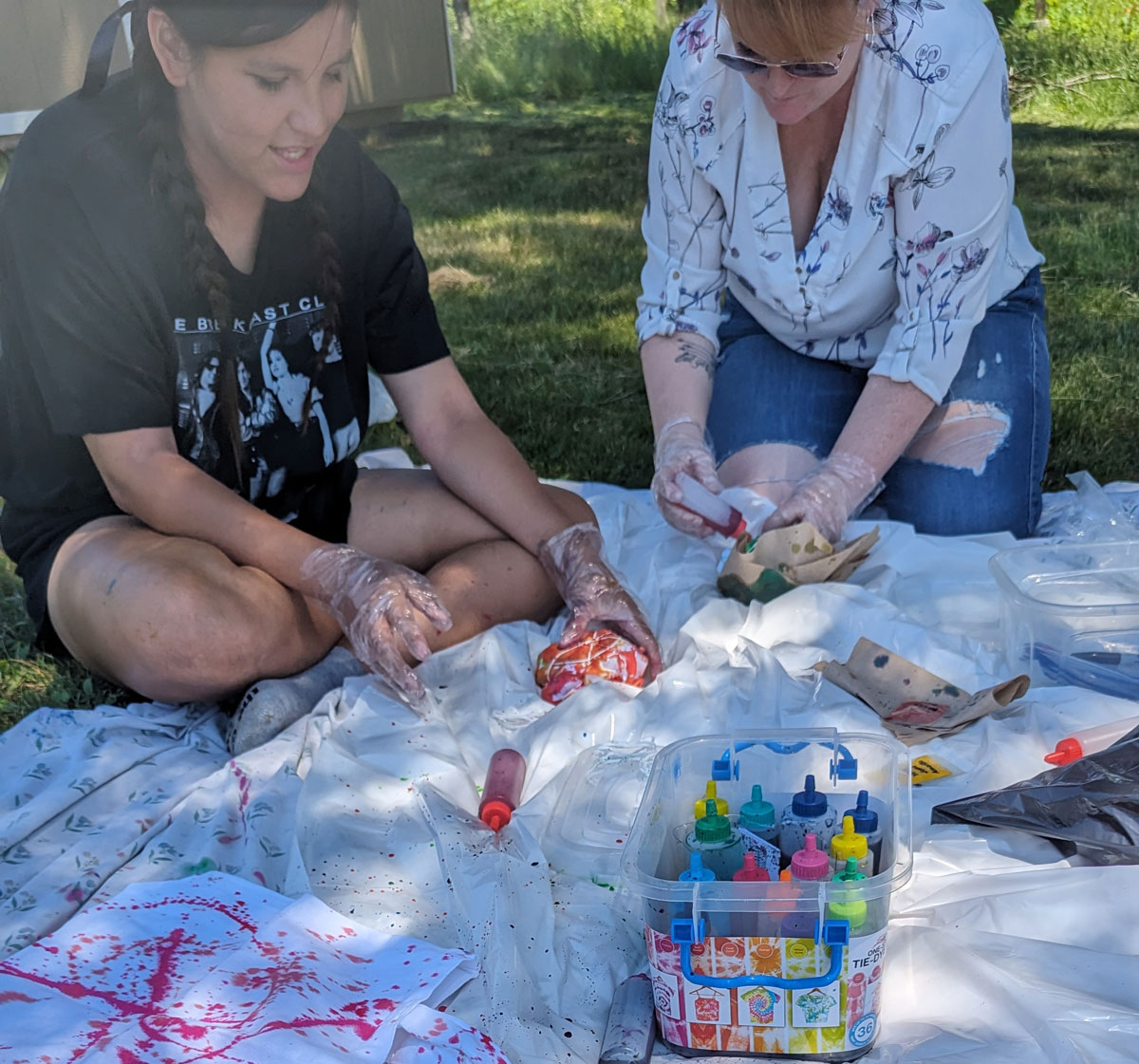 A teen in a black shirt with dark braided hair and a woman in a white shirt wearing glasses are making a craft with paint outside on the grass.