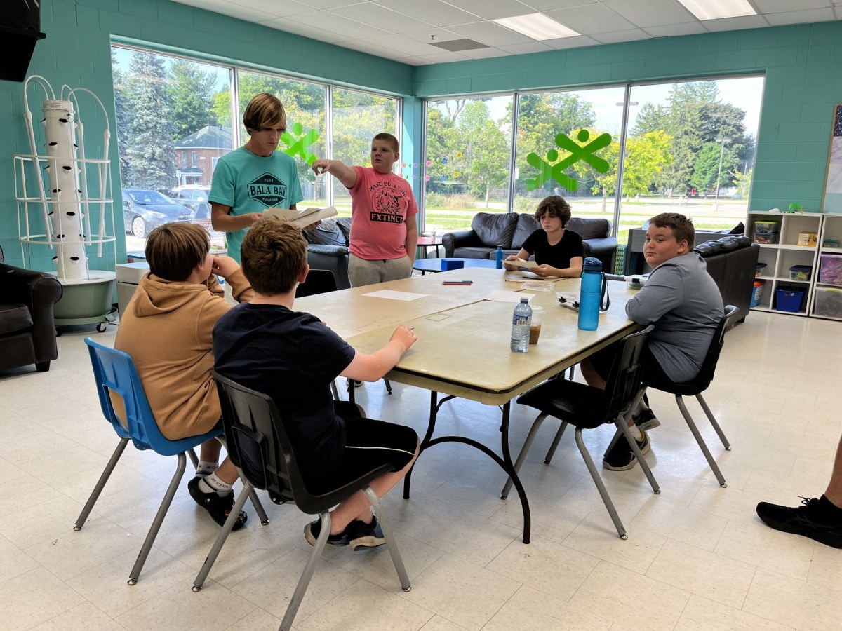 Four teen boys sit at a table as a man and another boy stand beside it speaking with them.