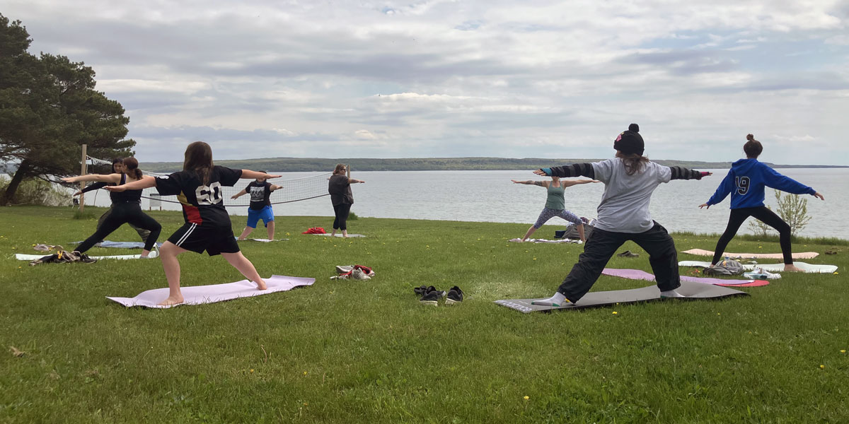 Teens standing on mats doing yoga poses on the grass beside a lake