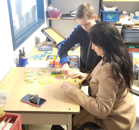 A woman is sitting at a table while a boy stands beside her. They are playing a game with yellow cards.