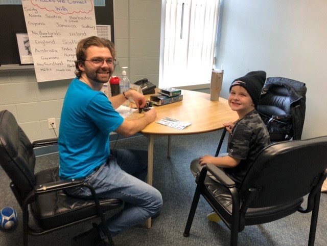 A young man in a blue shirt sits at a table with a smiling boy in a black hat.