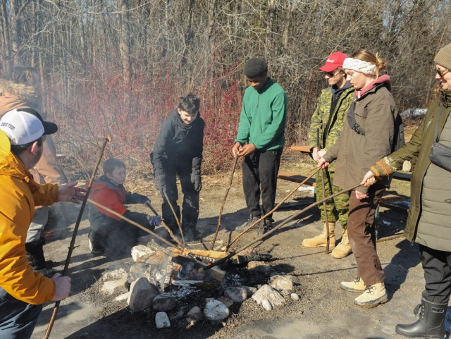 Adults, teens and a boy are standing around an open fire roasting marshmallows on long wooden sticks.