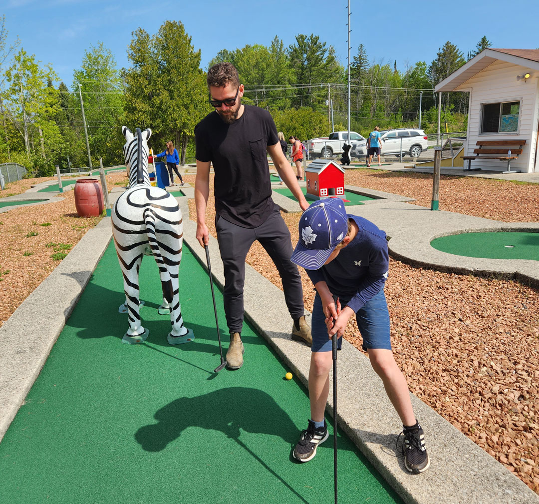 A man and young boy at a mini putt whole with a Zebra on it. The boy is about to putt.