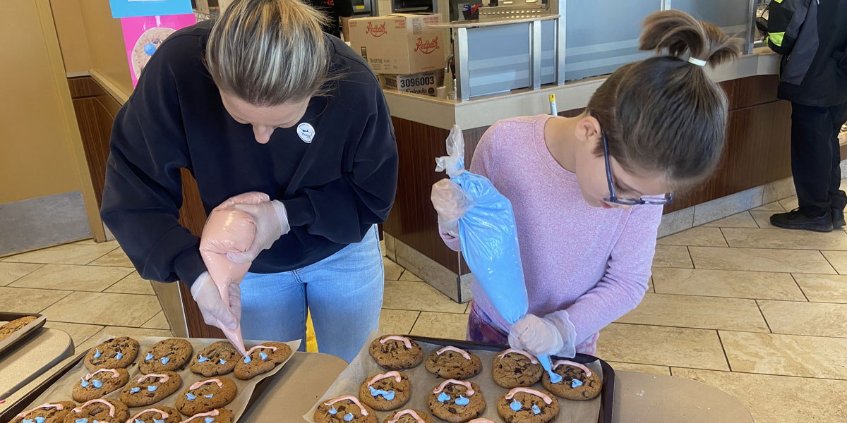 A woman and a young girl are putting smiles on chocolate chip cookies with icing bags