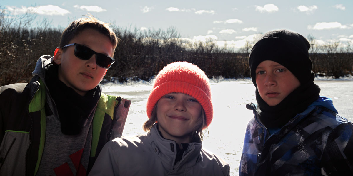 Three boys wearing winter clothes stand in front of a snowy lake